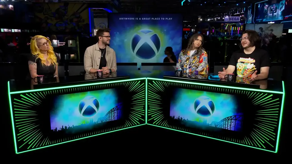 Четверо ведущих на фестивале gamescom обсуждают Xbox перед экраном с логотипом консоли и слоганом Anywhere is a Great Place to Play